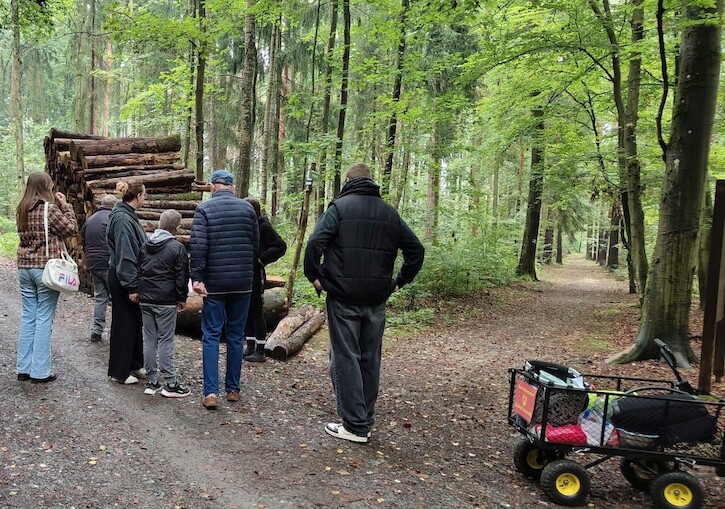 Die Schülerinnen, Schüler und Teilnehmer stehen an eine Weggabelung vor einem Holzhaufen.