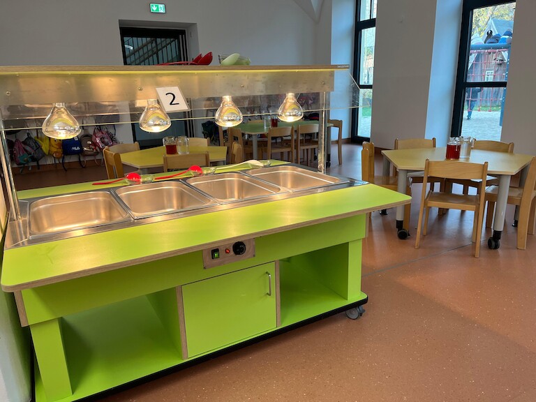 Ein Kindertisch im Hintergrund. Im Vordergrund eine Essenswagen, wo sich die Kinder die Speisen nehmen können.
