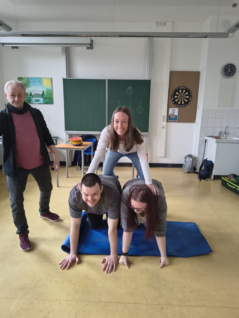 Zwei Schüler knien am Boden. Eine Schüler kniet darauf.