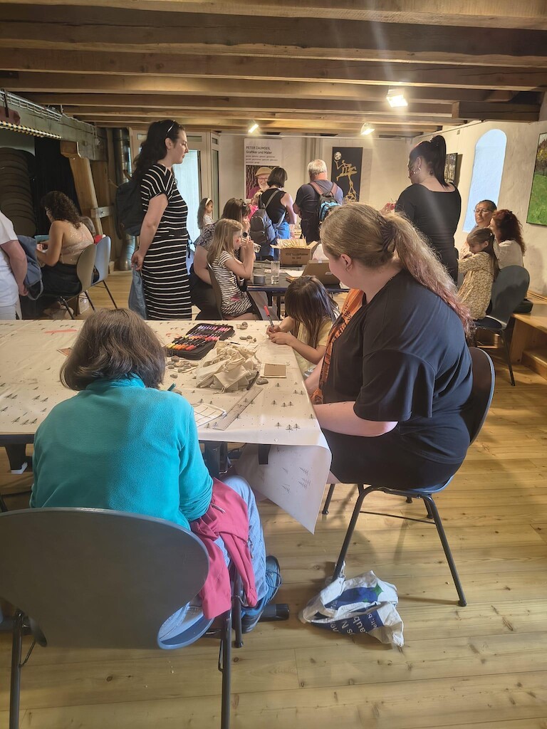 Im Raum sind mehrere Personen verteilt. Im Vordergrund sitzen zwei Kinder mit einer Schüler am Tisch.